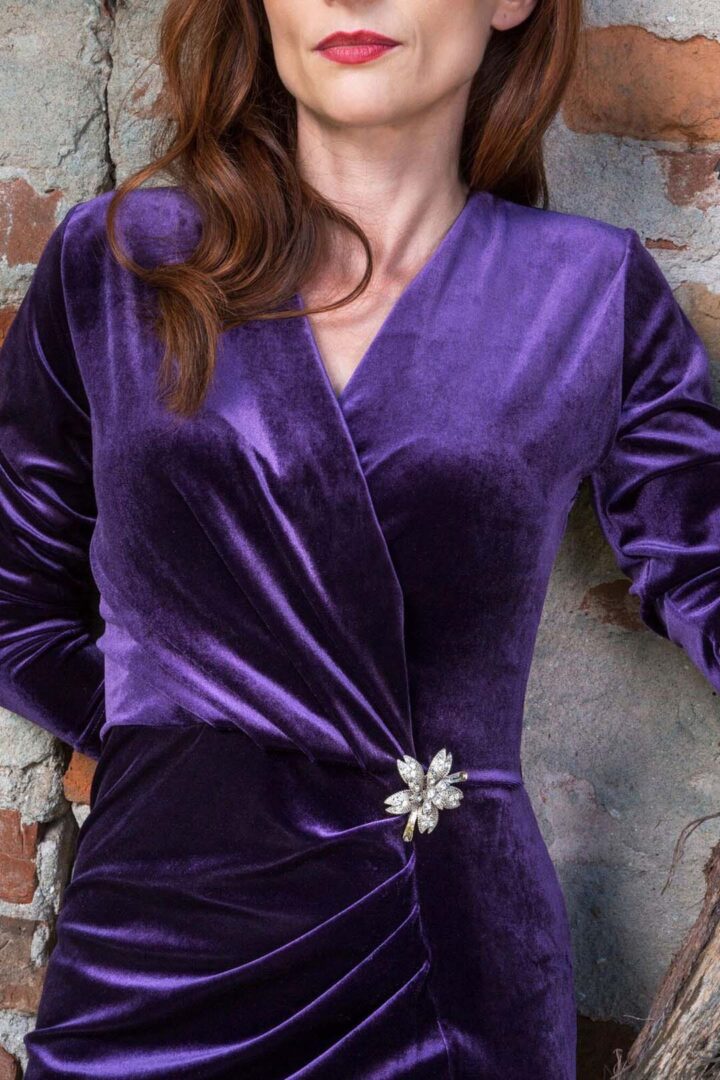 A woman in a purple velvet dress leaning against a brick wall.