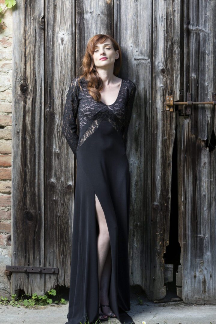 A woman in a black dress leaning against a wooden door.