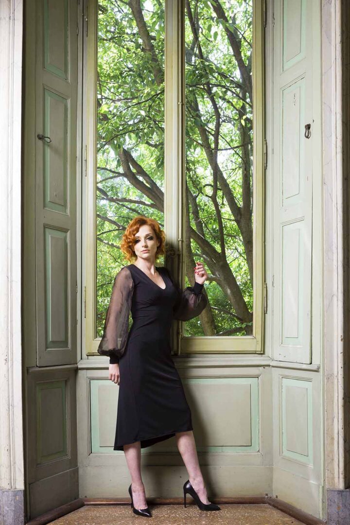 A woman in a black dress posing in front of a window.