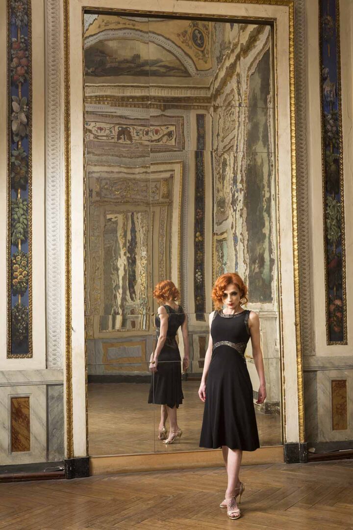 A woman in a black dress standing in front of an ornate mirror.