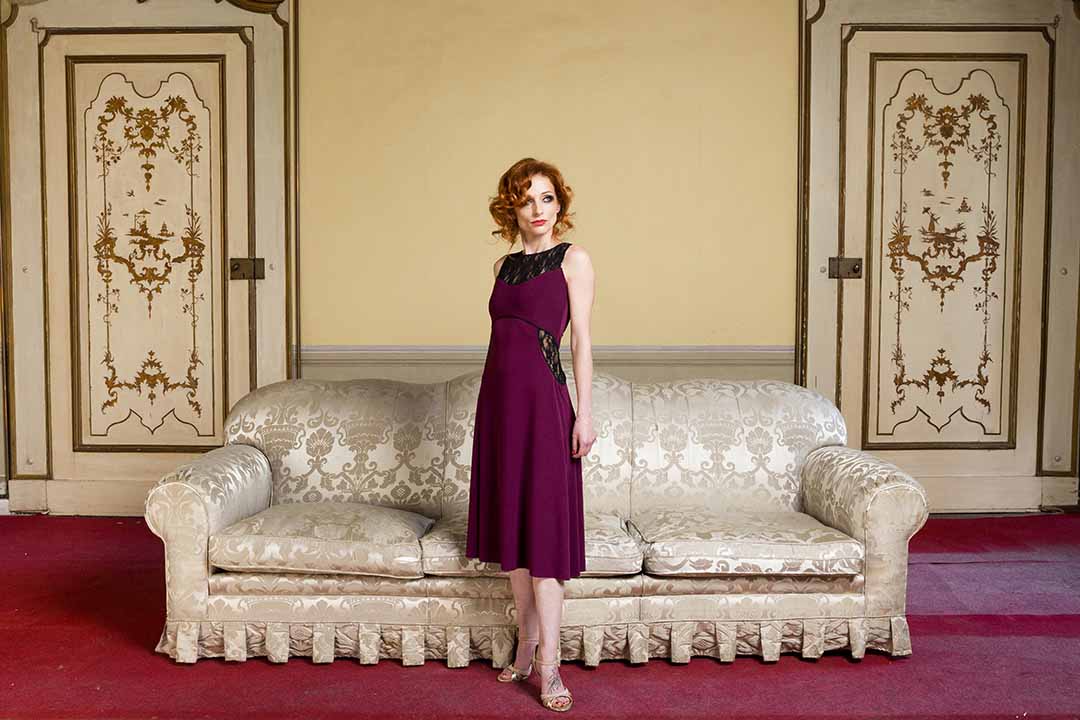 A woman in a purple dress standing in front of an ornate couch.