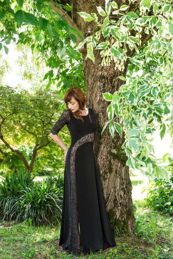 A woman in a black dress leaning against a tree.